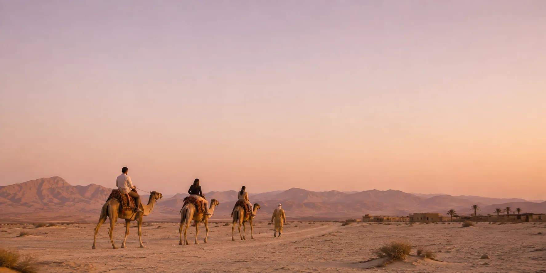 Three riders on camels crossing the desert at sunset, a handler walking alongside